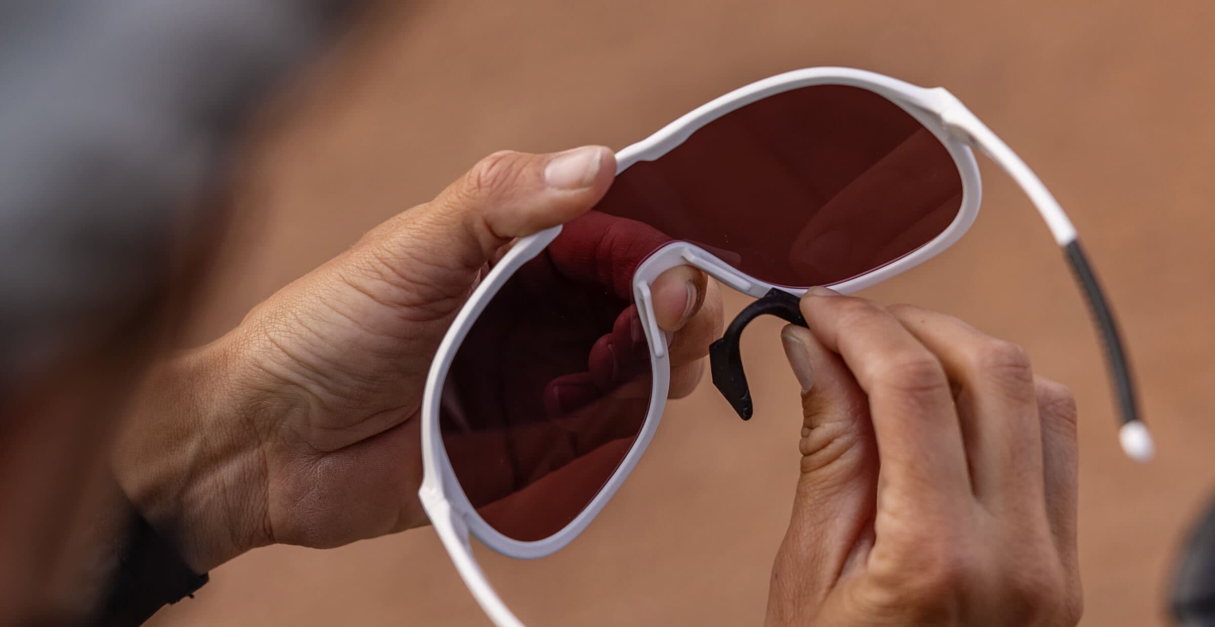 Two hands fitting a black nose pad to white ROKA sunglasses with a rose lens, red rock blurred behind