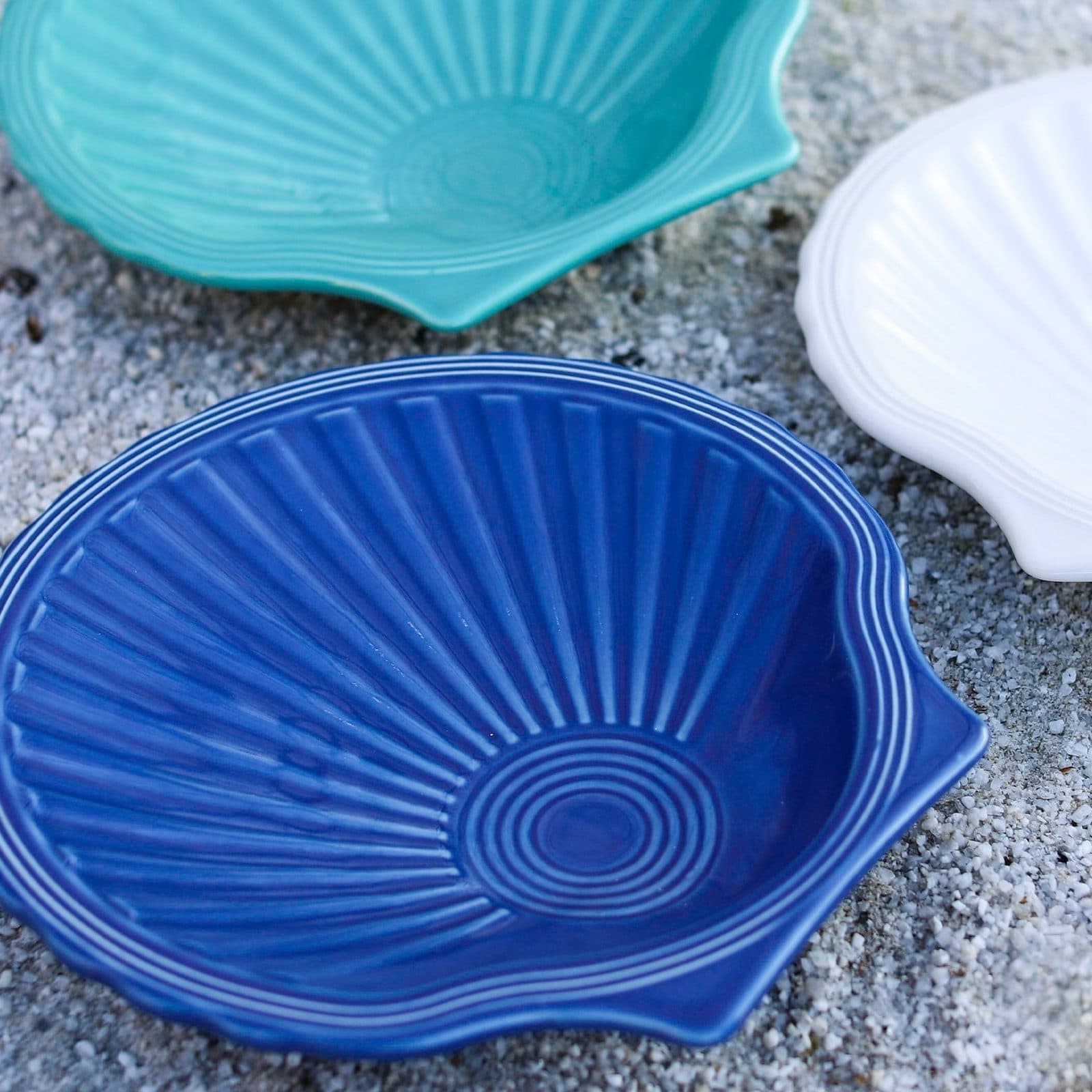 Three shell shaped plates, cobalt in front and turquoise and white behind, resting on sand