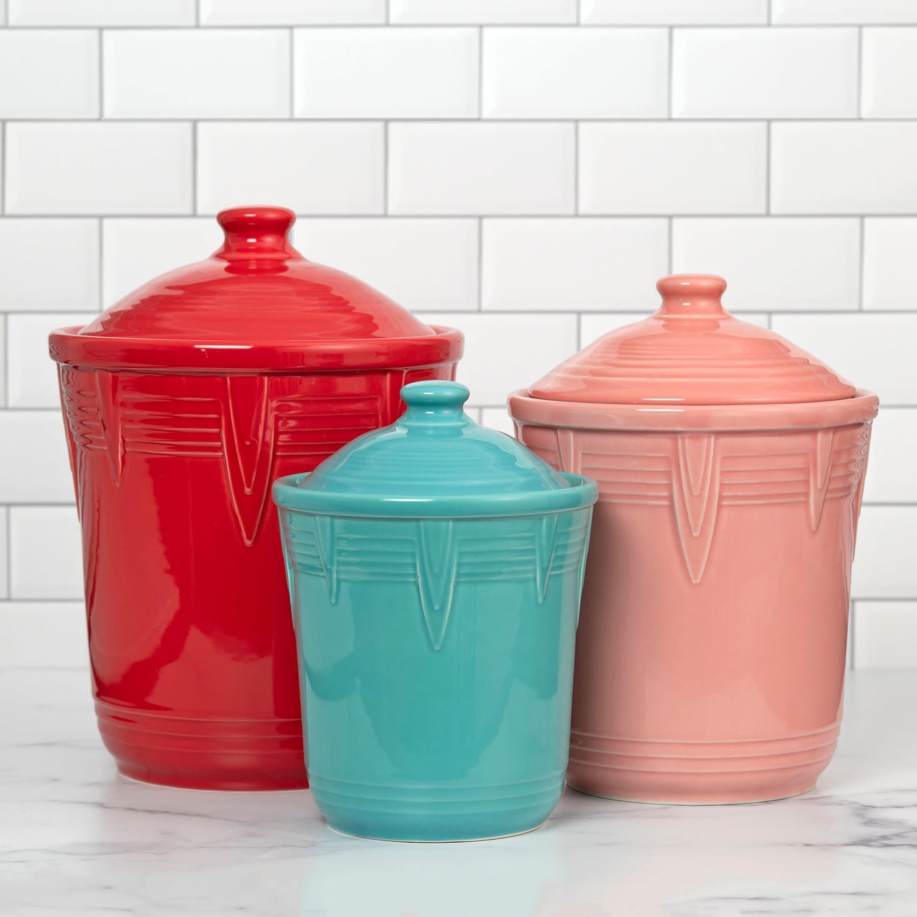 Three Fiesta chevron canisters in scarlet, turquoise and flamingo pink, in three sizes, on white marble against white subway tile.