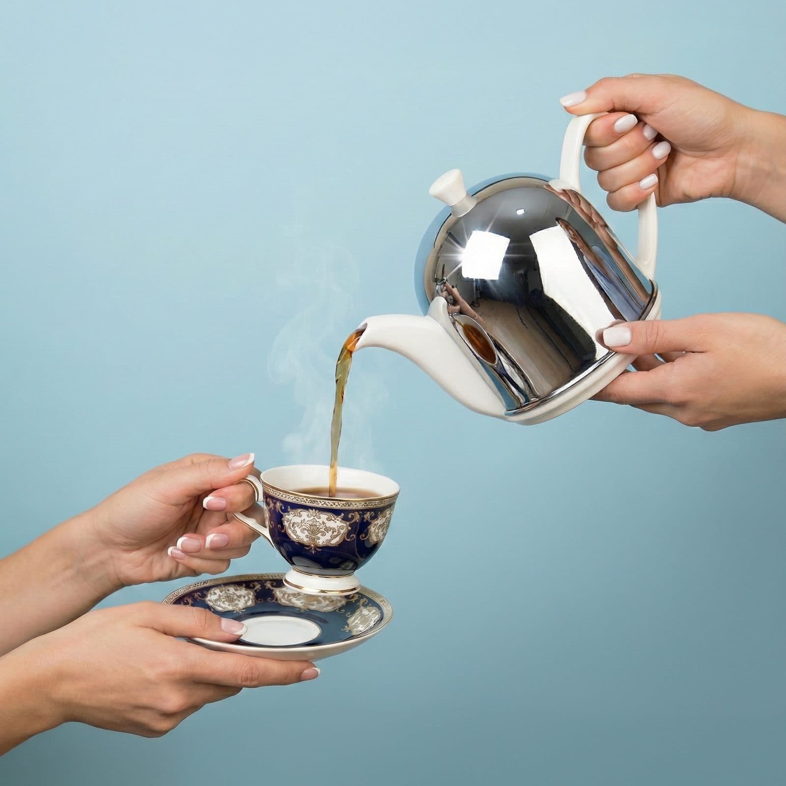 One hand pouring tea from a silver pot into a gilded blue teacup held out by another hand.
