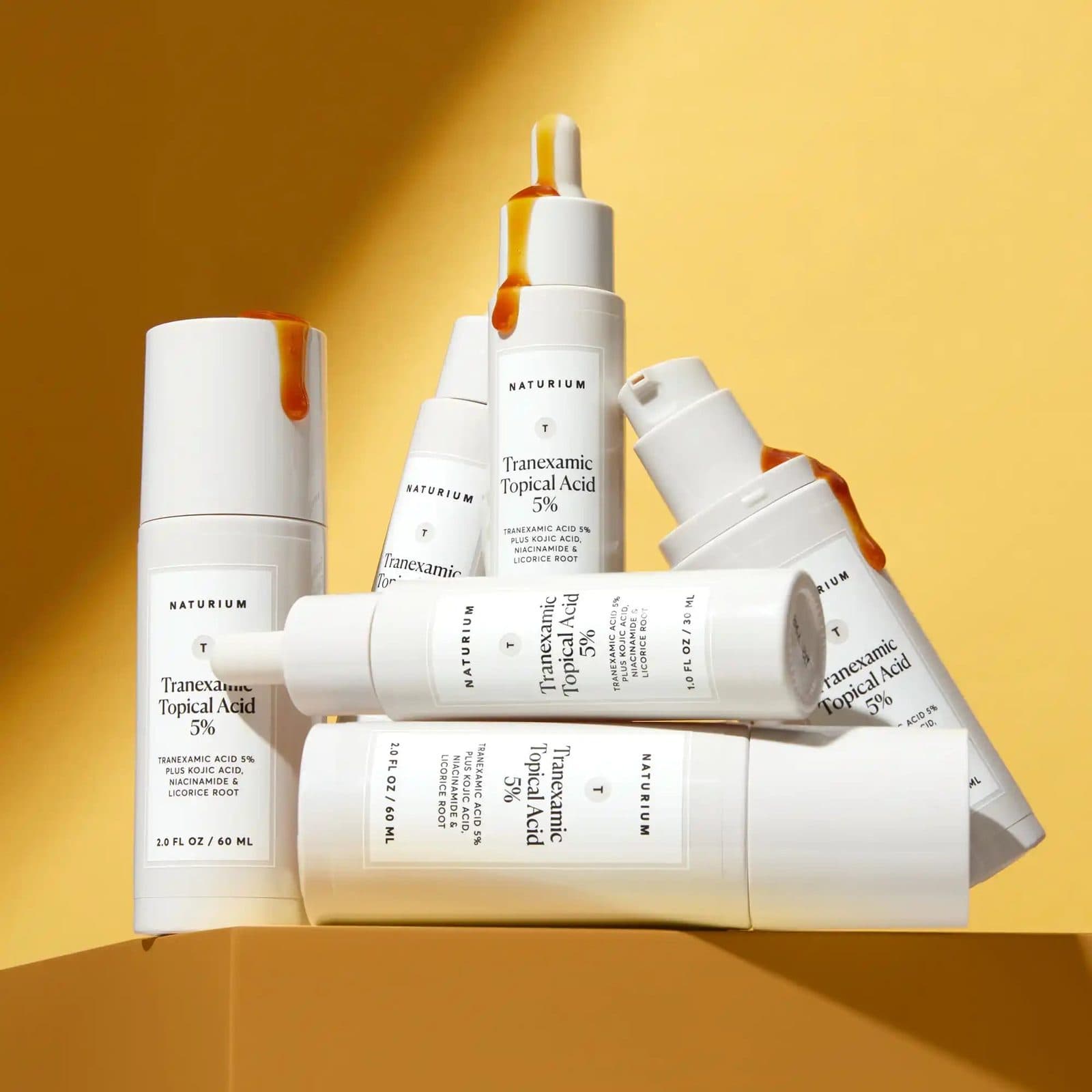 Six white serum bottles with amber drips stacked on a mustard block against a warm yellow wall.
