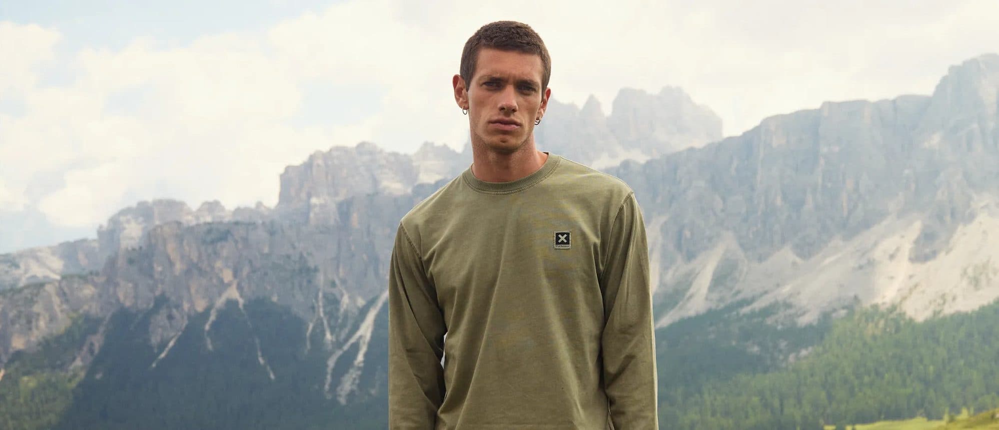 A man in an olive Blue Banana top on a green slope below jagged grey mountains.