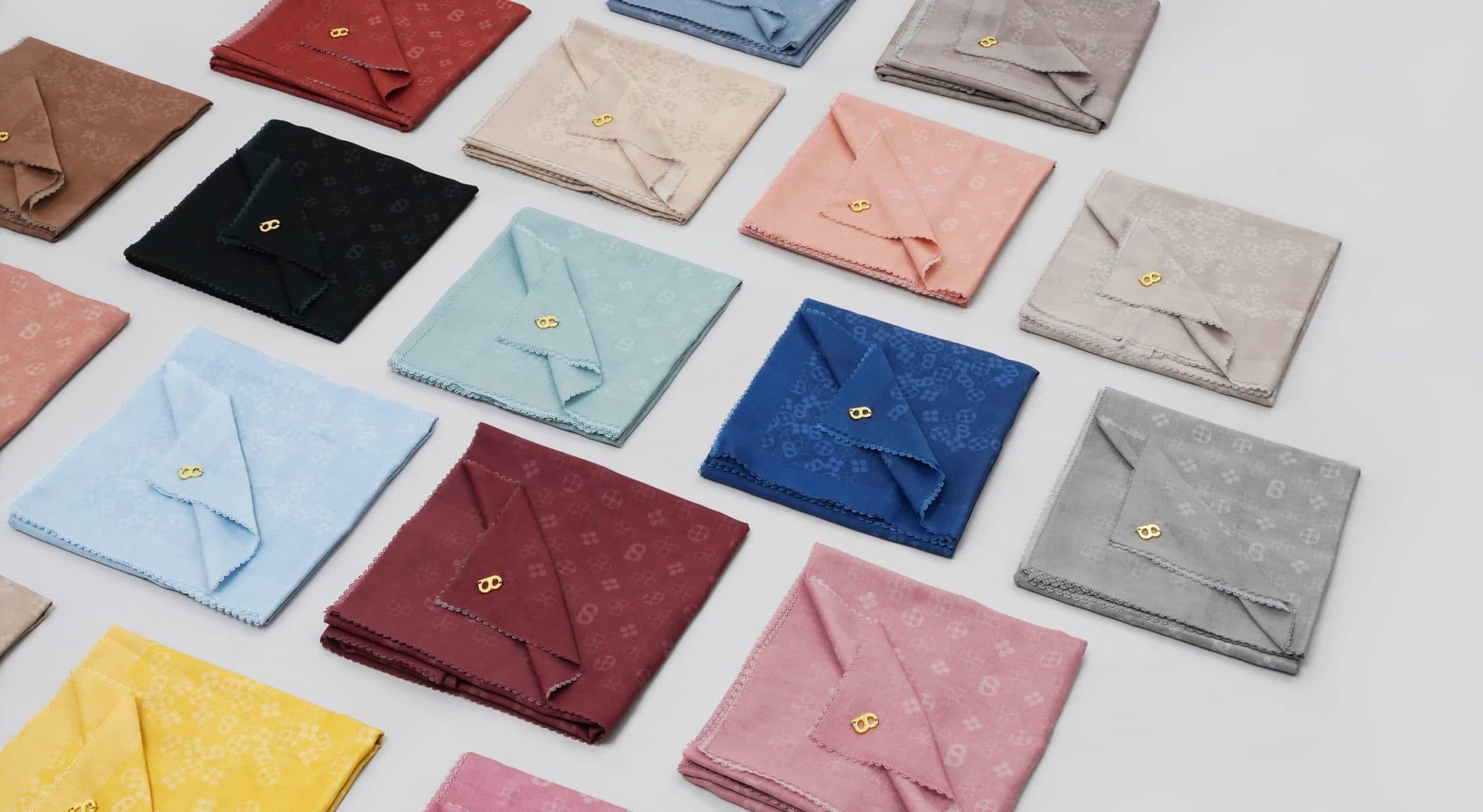 Monogram jacquard scarves folded into squares and laid out in rows on a pale grey surface, in black, burgundy, royal blue, sky blue, mint, blush, grey and yellow, each with a small gold pin.