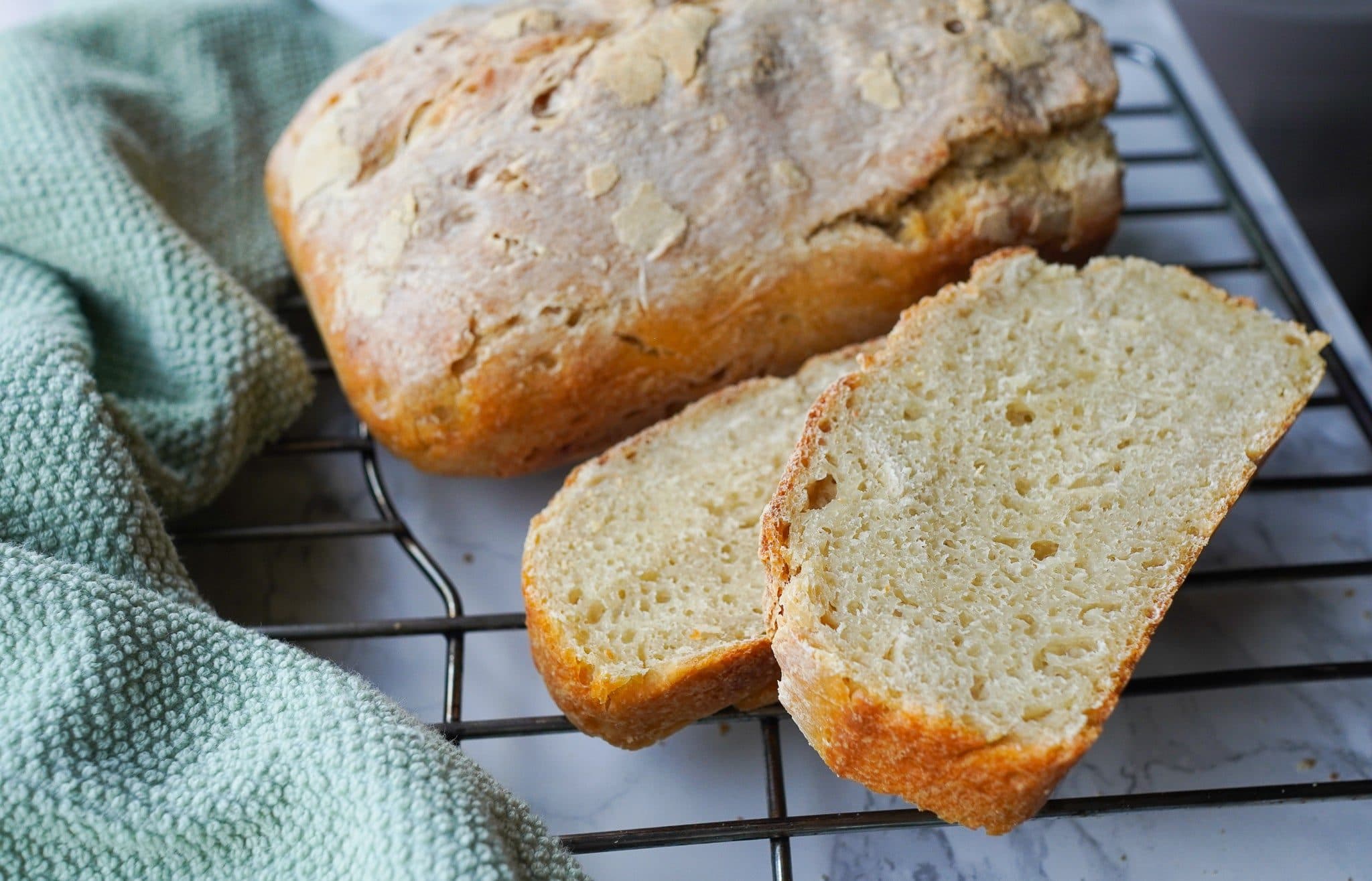 A homemade sourdough loaf and two thick slices on a black cooling rack over white marble, a sage green towel at the left.