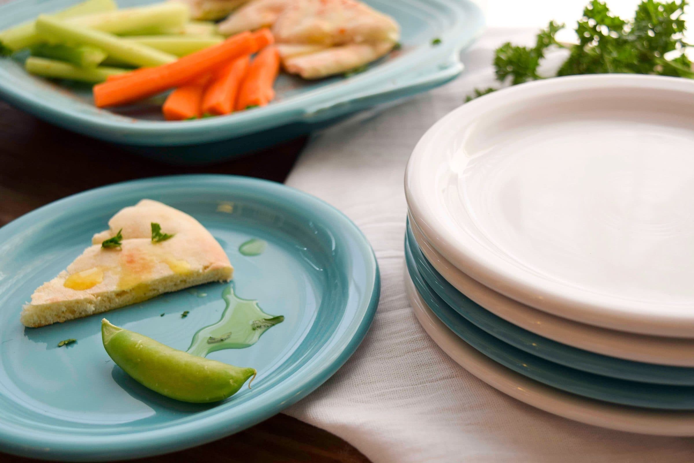 A turquoise Fiesta plate with a pita wedge and a snap pea on a white linen runner, white and turquoise plates stacked beside it, carrot and cucumber sticks on a platter behind.