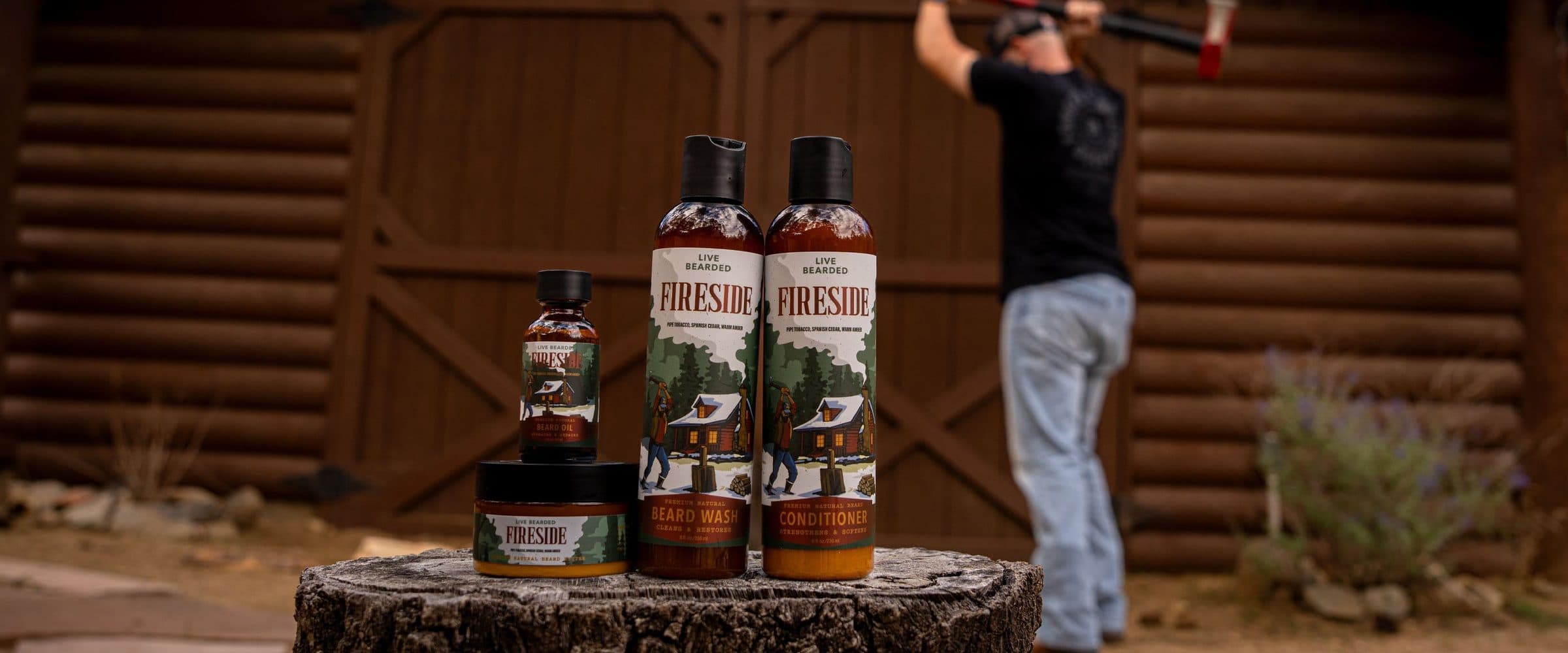 Live Bearded Fireside beard oil, butter, wash and conditioner on a tree stump outside a log cabin, a man splitting wood out of focus behind.
