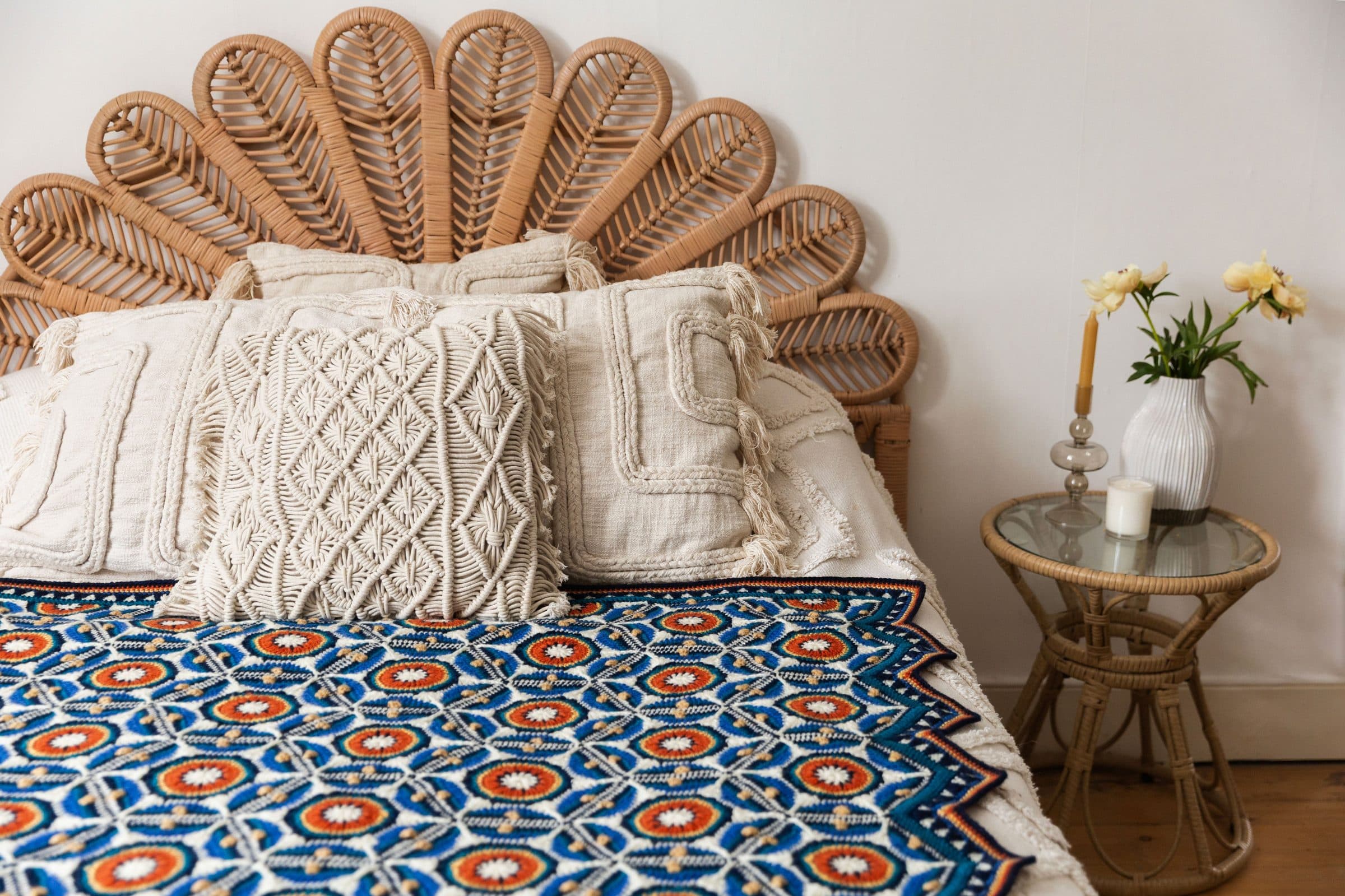 A Mary Maxim Sevilla crochet blanket in blue, white and orange hexagons across a bed with cream macrame pillows, a rattan side table with yellow flowers beside it.