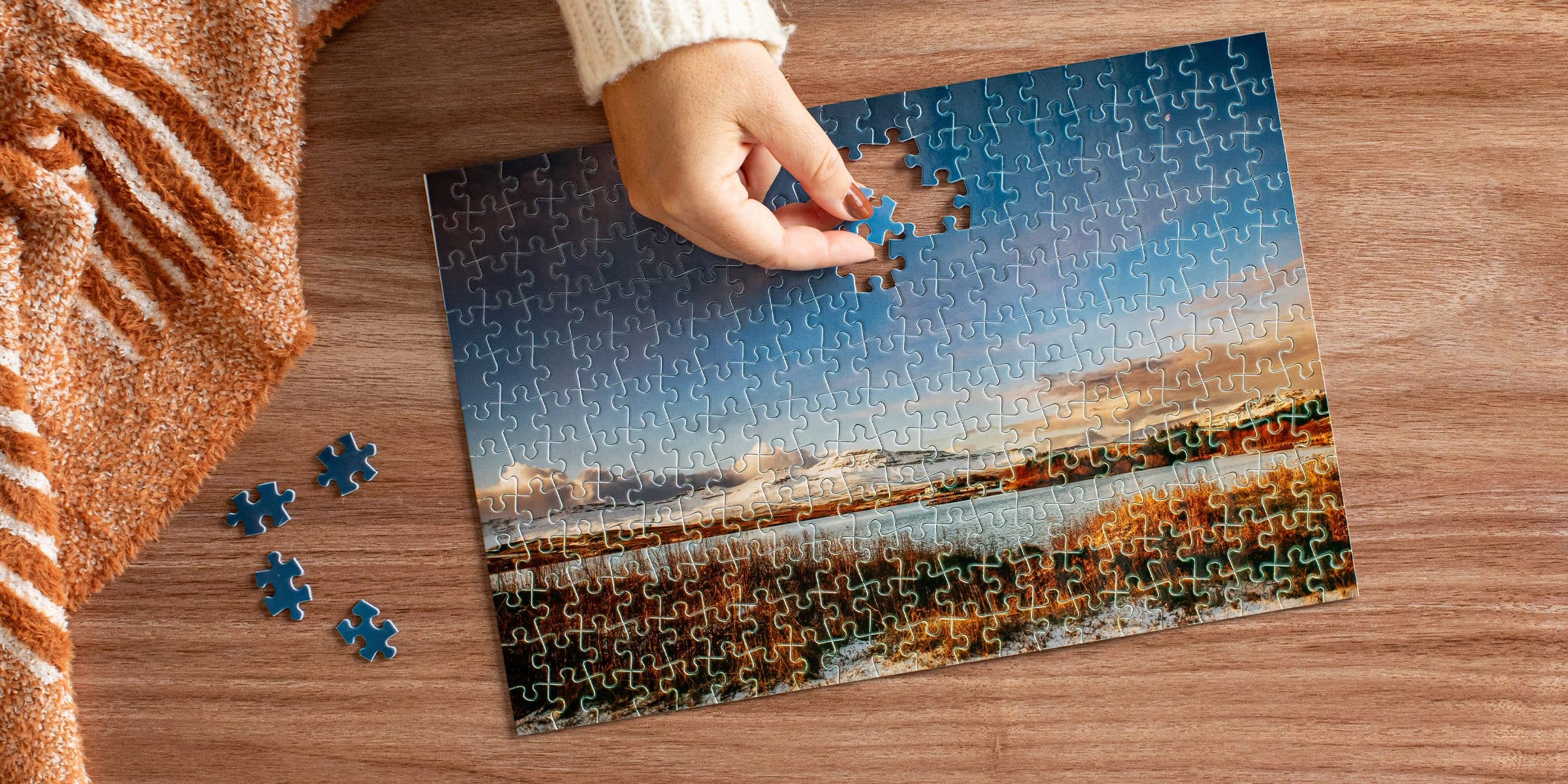 A hand fitting a piece into an Mpix photo puzzle of snowy mountains over golden grass, loose pieces and a striped throw on a wood table.