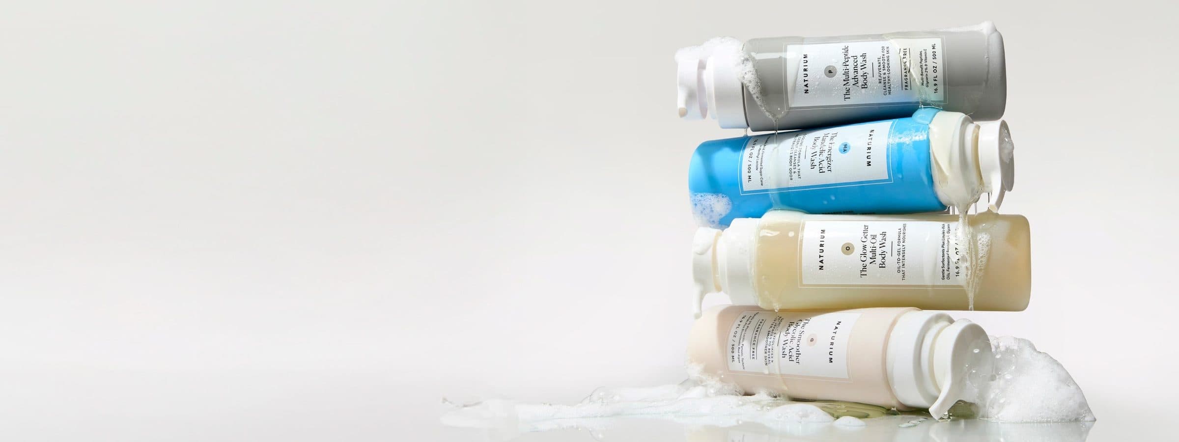 Naturium body washes stacked on their sides in soft lather on a grey backdrop.