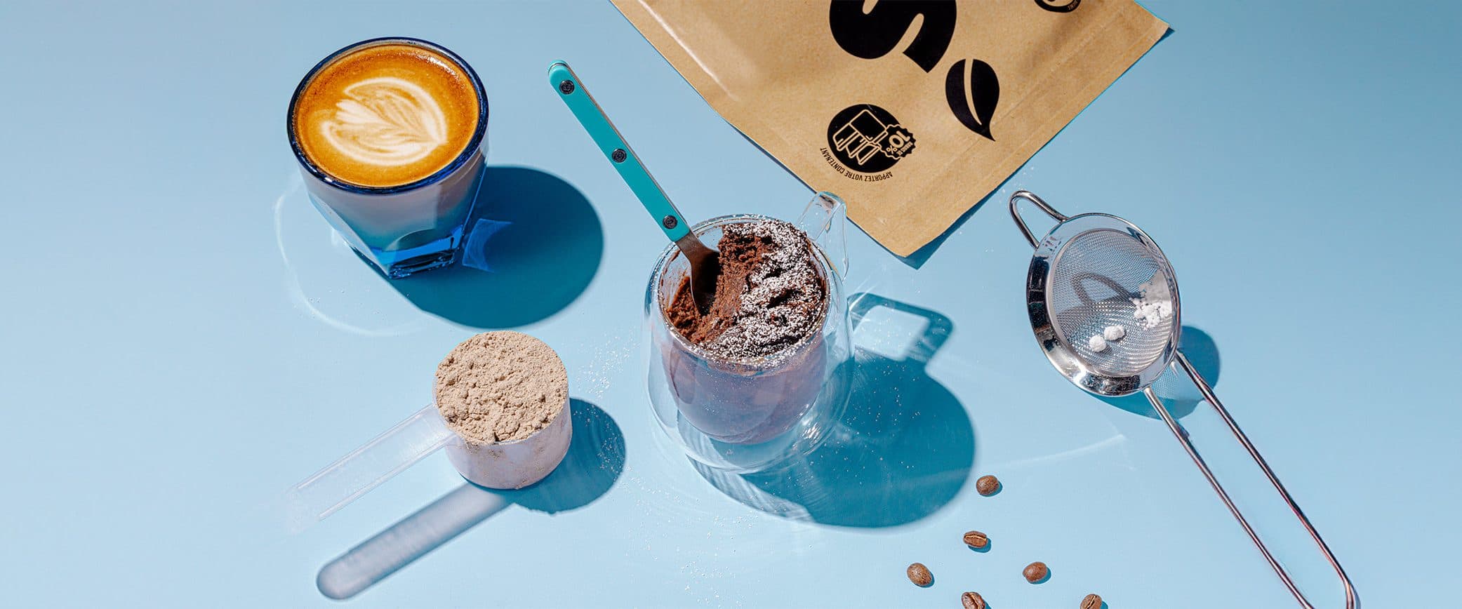 A scoop of chocolate protein powder, a glass mug cake dusted with icing sugar, a cortado, a small sieve and a few coffee beans on a pale blue table in hard sunlight, a Shop Santé kraft bag at the top.