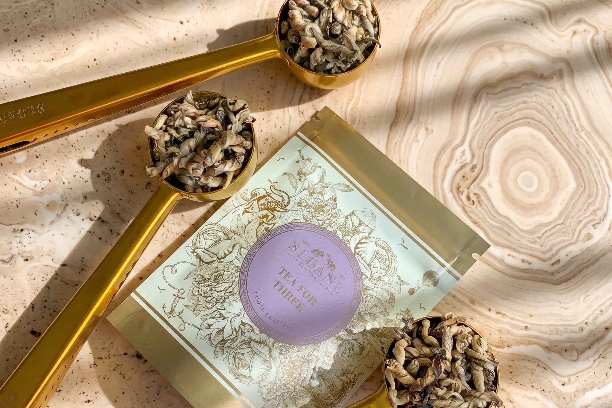 Three gold Sloane Tea scoops heaped with twisted loose leaf tea around a Sloane Tea for Three pouch, on cream stone streaked with leaf shadows.