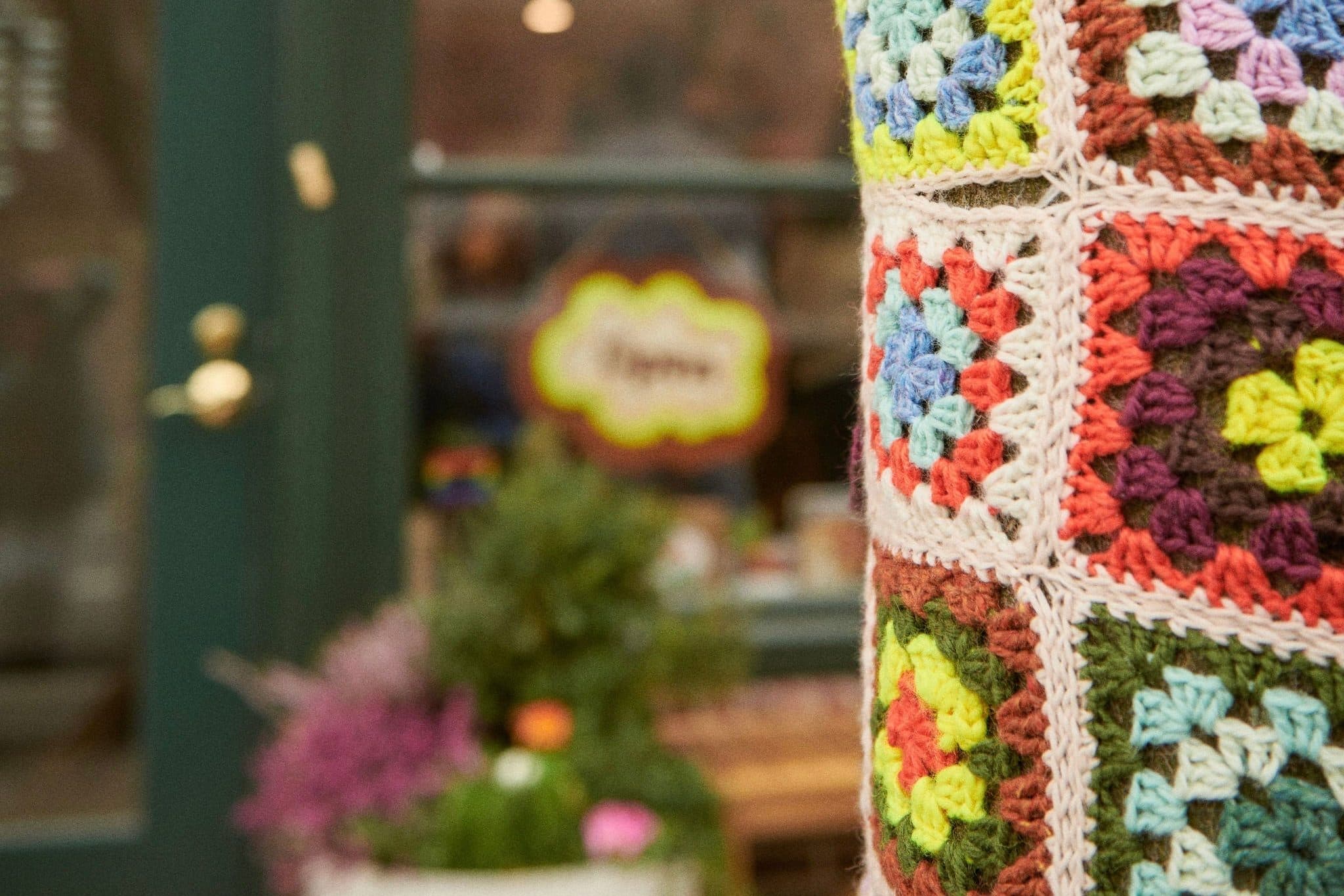 A crocheted cover of bright granny squares wrapped around a post outside a shop, a green door and a planter of flowers soft behind it.