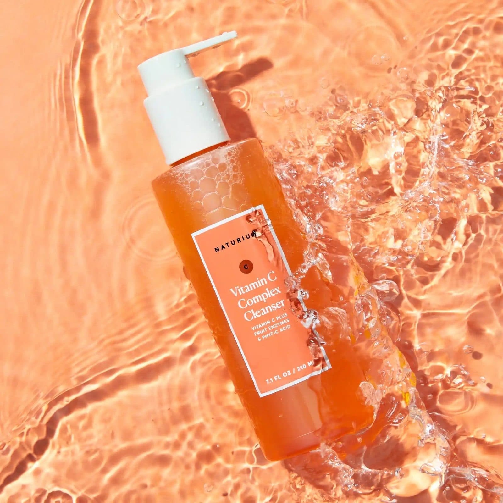 An orange cleanser bottle lying in rippling peach water, splashes around its pump.